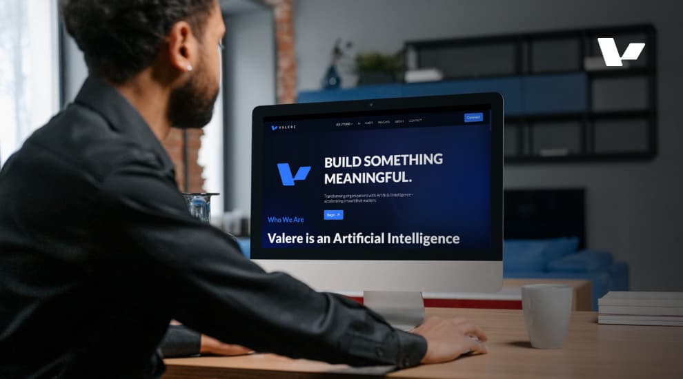 A man sitting at a desk, looking at a computer screen displaying Valere's website with the tagline 'Build Something Meaningful.' The screen highlights Valere as an Artificial Intelligence company.