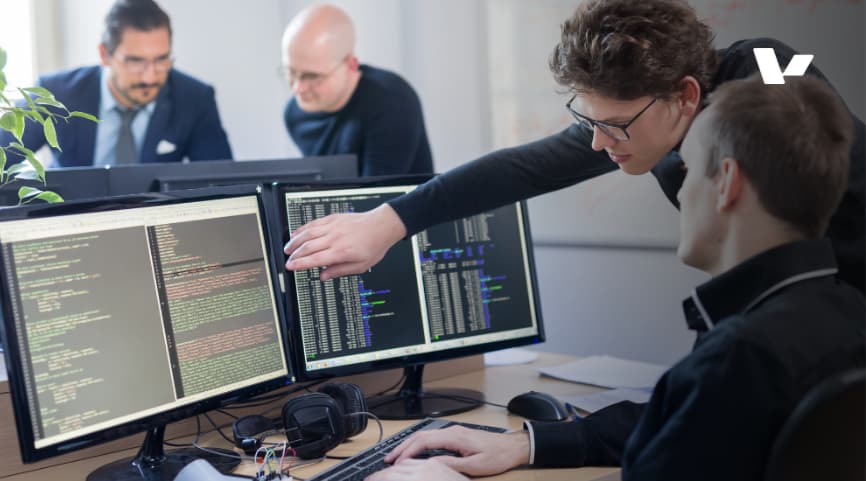 Two software engineers collaborating in an office, reviewing code on multiple computer screens during a team meeting, with one pointing at the code on the screen to provide guidance.