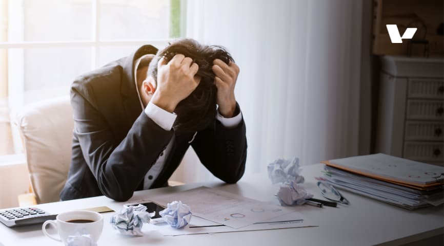 A stressed businessman sitting at his desk with his hands on his head, surrounded by crumpled papers, a coffee cup, and financial documents, reflecting frustration or burnout.