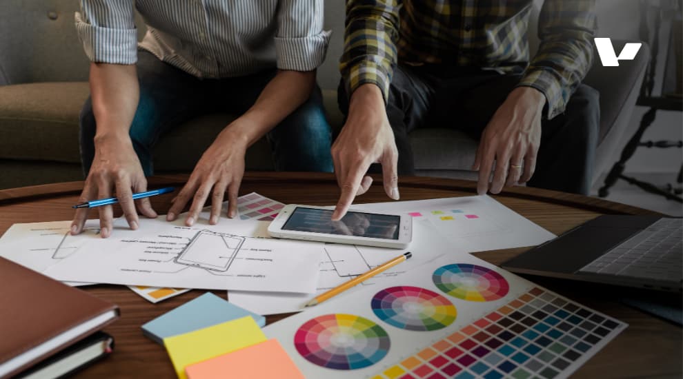 Two designers collaborating on a digital product design, with sketches, color palettes, and wireframes on the table, illustrating the early stages of UI/UX design development.