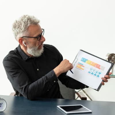 A professional older man with glasses and earphones points to a printed chart or graph in a folder while seated at a desk, explaining or presenting data during a meeting.