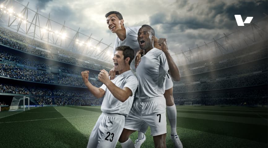 Three soccer players in white uniforms celebrate enthusiastically in a packed stadium, with bright lights and a cloudy sky.