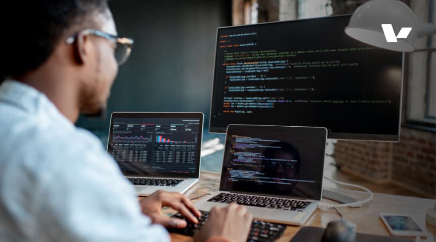"A software developer wearing glasses works on multiple screens displaying code and data analytics.