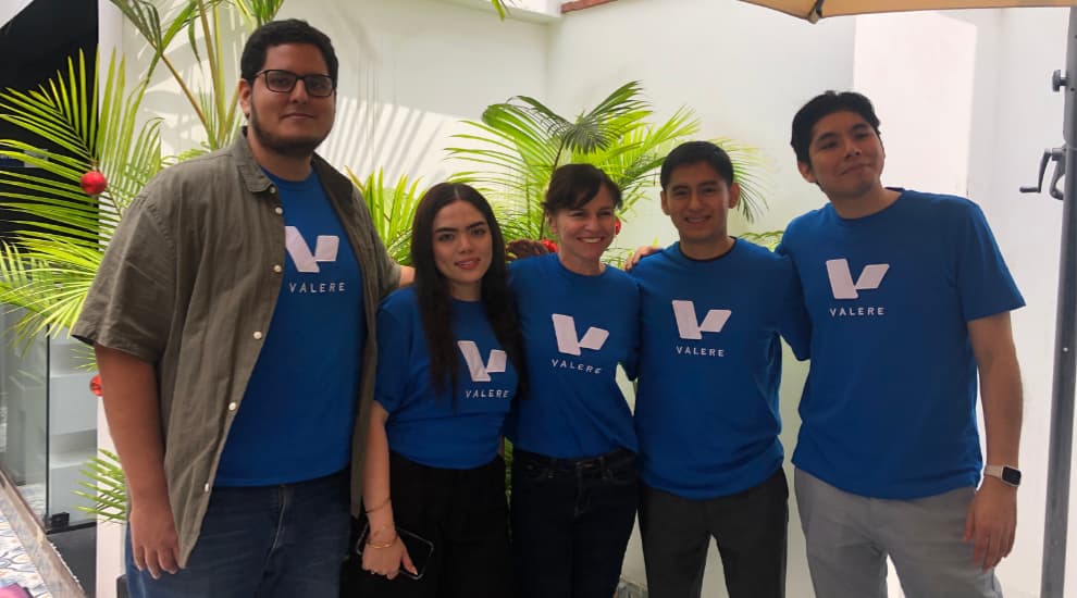 A group of five team members wearing blue 'Valere' branded t-shirts, standing together outdoors with tropical plants in the background, smiling and posing for the photo.