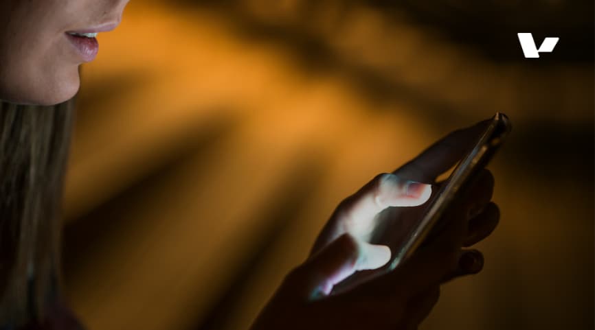 A close-up shot of a person using a smartphone in a dimly lit environment, with their finger gently tapping the screen. The focus is on the phone's glowing display and the person's hand.