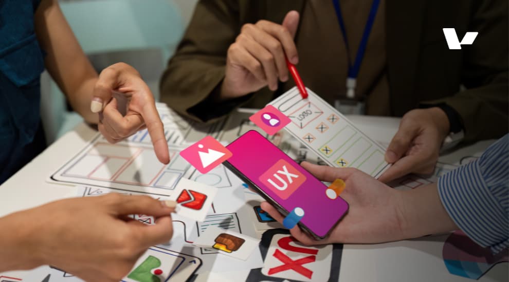 A collaborative team is working on a UX design project, with hands pointing at mobile app wireframes and colorful design elements on a table, emphasizing user experience and interface design.