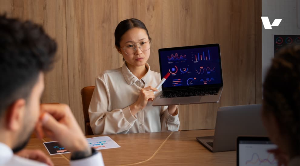 A woman in a business meeting presenting graphs and charts on a laptop screen to two colleagues, demonstrating data analysis in a professional setting.