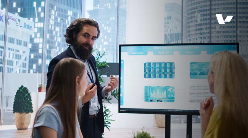 A business professional presenting data and statistics on a screen to two colleagues during a meeting in a modern office setting. The presentation includes graphs, charts, and information on performance metrics.